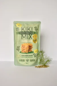 Package of Kiki Seed Cracker Mix, Fennel Seed flavor, standing upright with a small pile of fennel seeds and a fennel sprig beside it on a white background