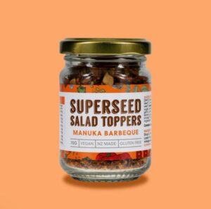 Glass jar of Superseed Salad Toppers in Manuka Barbeque flavour with a gold lid, shown against a orange background. The label highlights vegan, gluten-free, NZ made, and 70g, with visable crunchy seed and nut pieces inside the jar