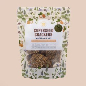 Cream-colored packet of Superseed Crackers in Macadamia Nut flavour. The packaging features illustrated herbs, flowers, and macadamia nuts, with a clear window showing seeded crackers inside. Front text includes ‘Superseed Crackers – Macadamia Nut,’ ‘Natural Ingredients,’ ‘Low Carbs,’ ‘Gluten Free,’ ‘Vegan,’ ‘Keto,’ and ‘Slow baked until crispy