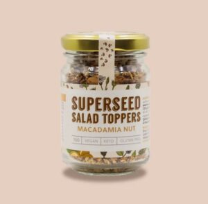 Glass jar of Superseed Salad Toppers in Macadamia Nut flavour with a gold lid, shown against a light neutral background. The label highlights vegan, keto, gluten-free, and 70g, with visible chopped nuts and seeds inside the jar