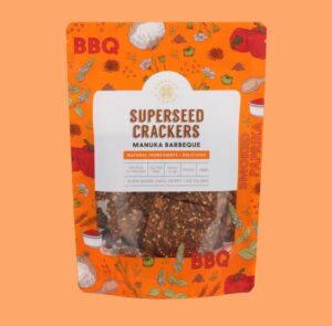 Bright orange packet of Superseed Crackers in Manuka Barbeque flavour. The packaging features illustrated vegetables and spices, with a clear window showing seeded crackers inside. Text on the front reads ‘Superseed Crackers – Manuka Barbeque,’ ‘Natural Ingredients,’ ‘Gluten Free,’ ‘Vegan,’ and ‘Slow baked until crispy